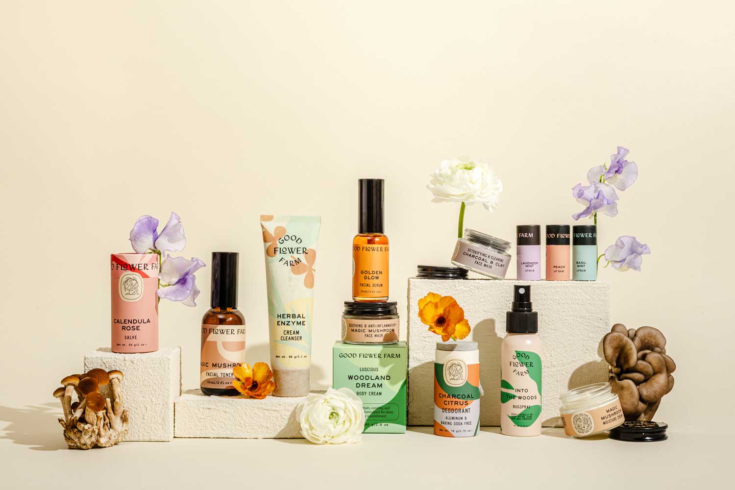 A variety of skincare and wellness products are arranged on white blocks, surrounded by white and purple flowers and mushrooms, against a beige background.