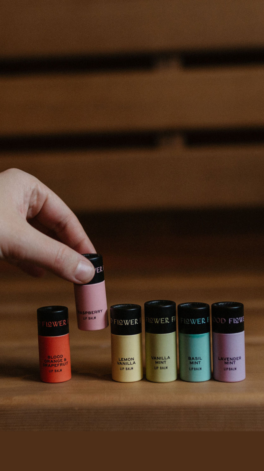 A hand picks a tube from the Good Flower Farm Rainbow Organic Lip Balm Set of 9, with nine colorful, moisturizing flavors displayed on a wooden surface against a blurred brown background.