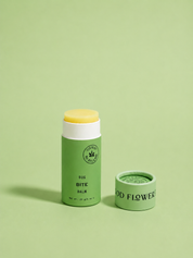 A green tube of Good Flower Farm Bug Bite Balm For Anti-Itch stands upright with its lid off on a matching green background, revealing the yellow balm inside. The lid is placed beside the tube.