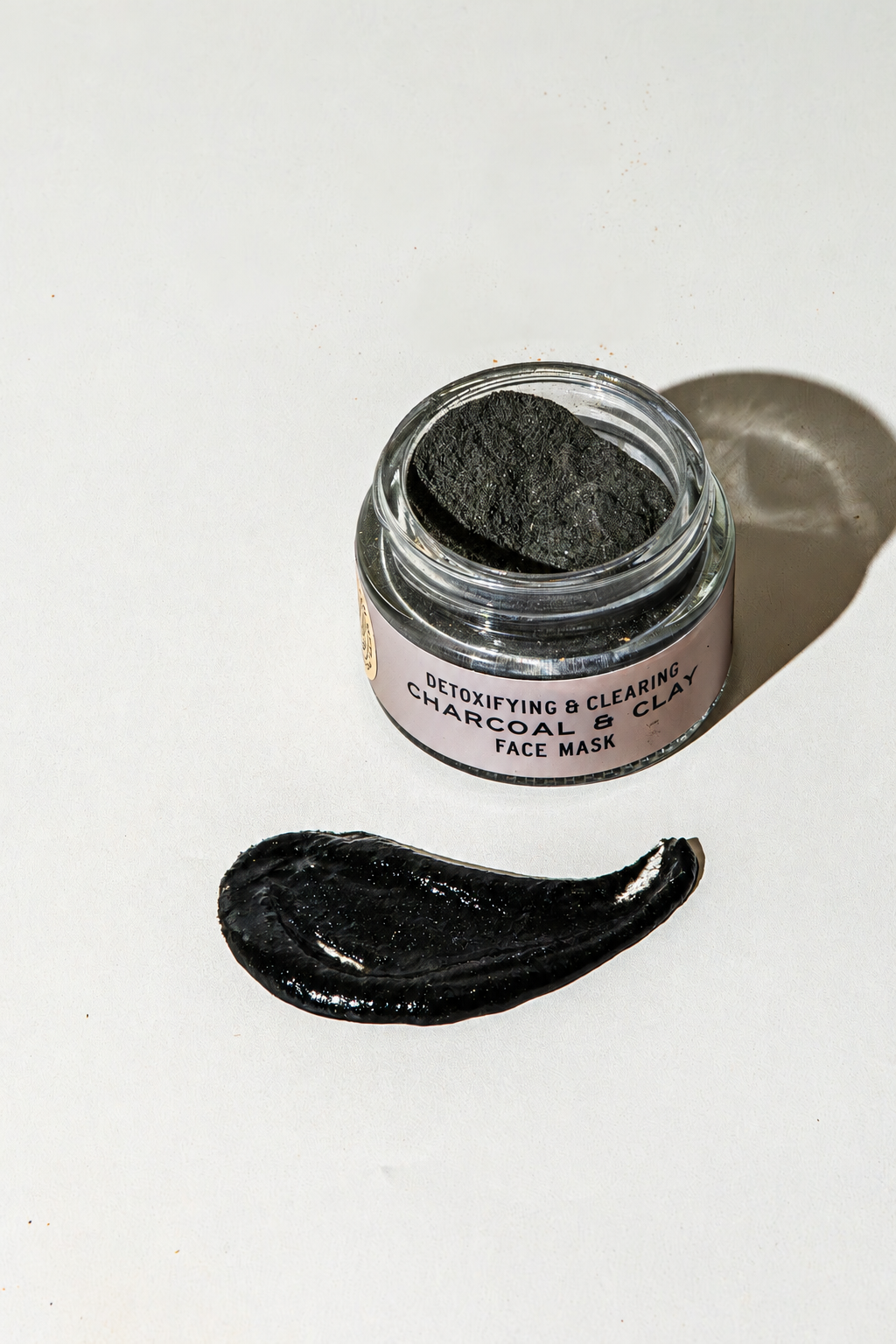 An open glass jar of Good Flower Farm Charcoal & Clay Botanical Face Mask next to a smear of the product on a white surface.