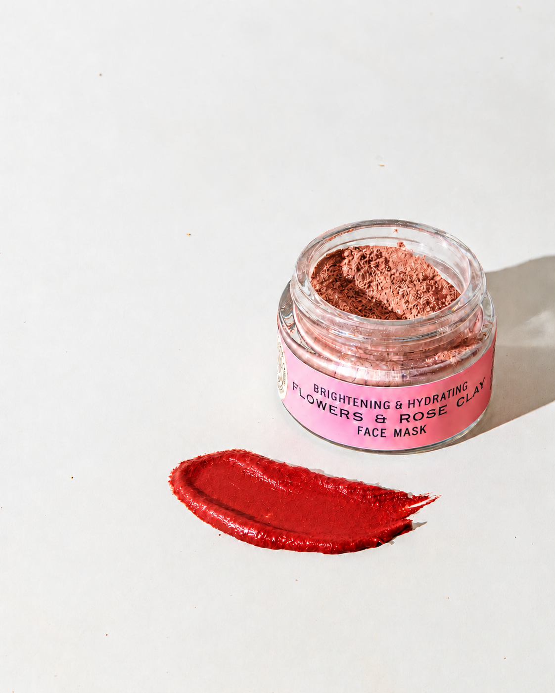 A glass jar of Good Flower Farm Flowers & Rose Clay Botanical Face Mask with a scoop of smooth, deep red product swiped on the surface below it.