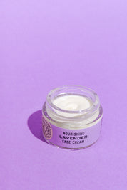 A glass jar of Good Flower Farm’s Lavender Face Cream sits open on a purple surface, revealing the creamy white, calendula-infused moisturizer inside.