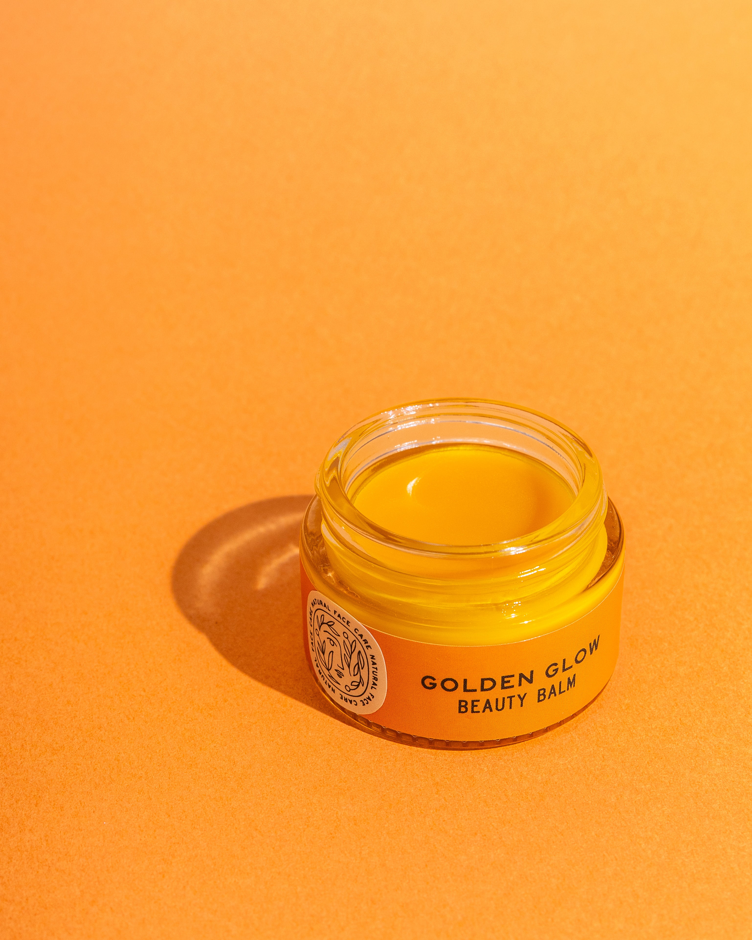 A small glass jar labeled “TESTER — Golden Glow Beauty Balm” from Good Flower Farm sits on an orange surface, casting a soft shadow. The open jar shows the smooth, vitamin C-infused yellow balm inside.
