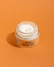 A small glass jar of Good Flower Farm’s Magic Mushroom Moisture Cream, with its lid off to display the creamy product inside, sits on an orange surface; the formula is infused with adaptogenic maitake.