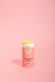 A tube of Good Flower Farm Calendula Rose Salve with a pink and cream label stands upright on a light pink background, the balm slightly exposed to showcase its soothing formula and eco-friendly packaging.