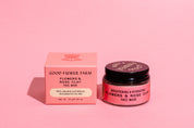A pink jar and matching box of Good Flower Farm’s Flowers & Rose Clay Botanical Face Mask sit on a pink surface. The open jar reveals the lid, and both items are labeled for brightening and hydrating benefits.