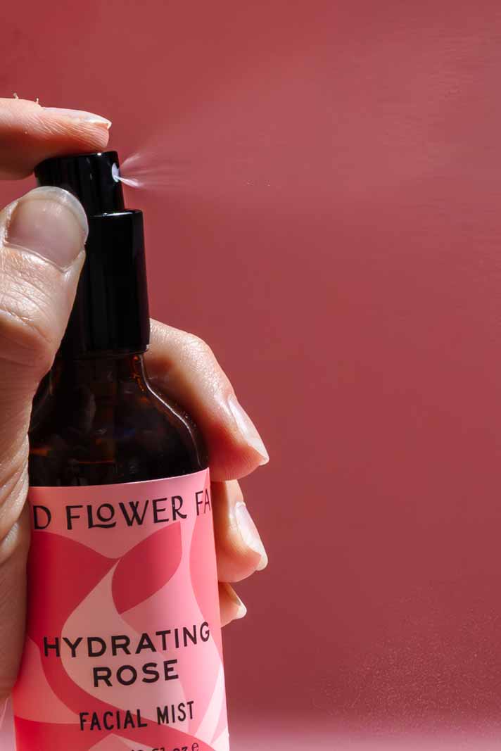 A hand sprays a bottle from the good-flower-farm Dry Skin Bundle: Save 15% against a pink background. Mist visibly streams from the nozzle, delivering a burst of hydration—ideal for moisturizing and soothing dry skin.