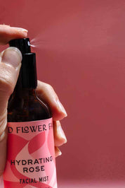 A hand sprays a bottle from the good-flower-farm Dry Skin Bundle: Save 15% against a pink background. Mist visibly streams from the nozzle, delivering a burst of hydration—ideal for moisturizing and soothing dry skin.