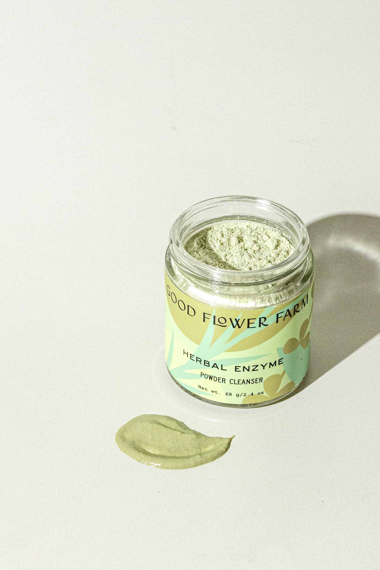 A jar of Good Flower Farm’s TESTER — Herbal Enzyme Powder Facial Cleanser rests on a light surface with green powder, formulated with coconut milk and aloe vera, displayed in front. The label features a pale green background with yellow and brown designs.