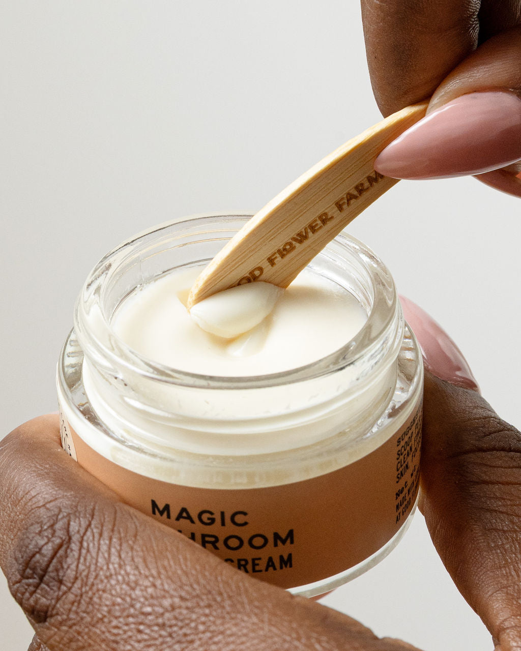 A hand holds a jar of cream as another uses a Good Flower Farm Reusable Bamboo Spatula (from the Set of 5) to scoop some out.