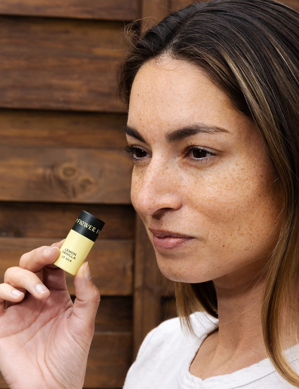 A woman with light brown hair and freckles holds a Good Flower Farm Lemon Vanilla Organic Lip Balm near her face, standing in front of a wooden background.