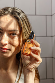 A woman with long brown hair holds up a bottle of Good Flower Farms Golden Glow Facial Serum in sunlight against a tiled wall, showcasing the serums promise of natural radiance and skin hydration.