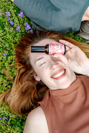 A smiling woman lies on grass with purple flowers, holding a pink bottle from the good-flower-farm Dry Skin Bundle: Save 15% over one eye, hinting at the secret to facial moisturizing and soft, radiant skin.