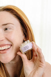 A smiling woman with long red hair holds a jar of Good Flower Farm’s Lavender Face Cream near her face, showing off its soothing and hydrating lavender formula.