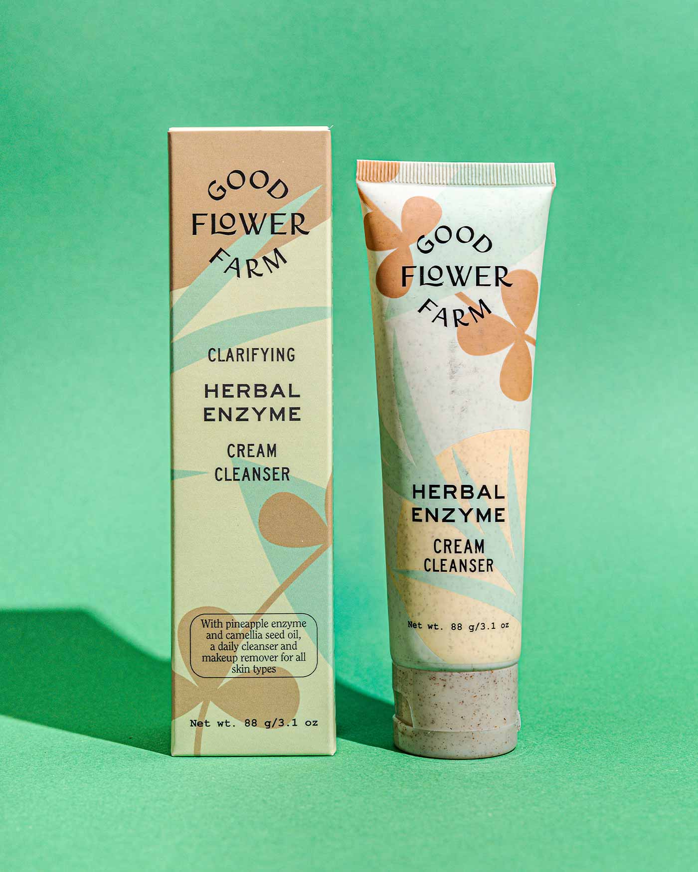 A tube and box of Good Flower Farm Herbal Enzyme Cream Cleanser &amp; Makeup Remover, featuring abstract leaf designs and product details, are displayed on a green background.