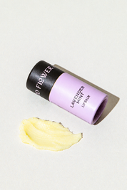 A lavender-colored cardboard tube of Good Flower Farm Lavender Mint Organic Lip Balm lying next to a smear of yellow balm.