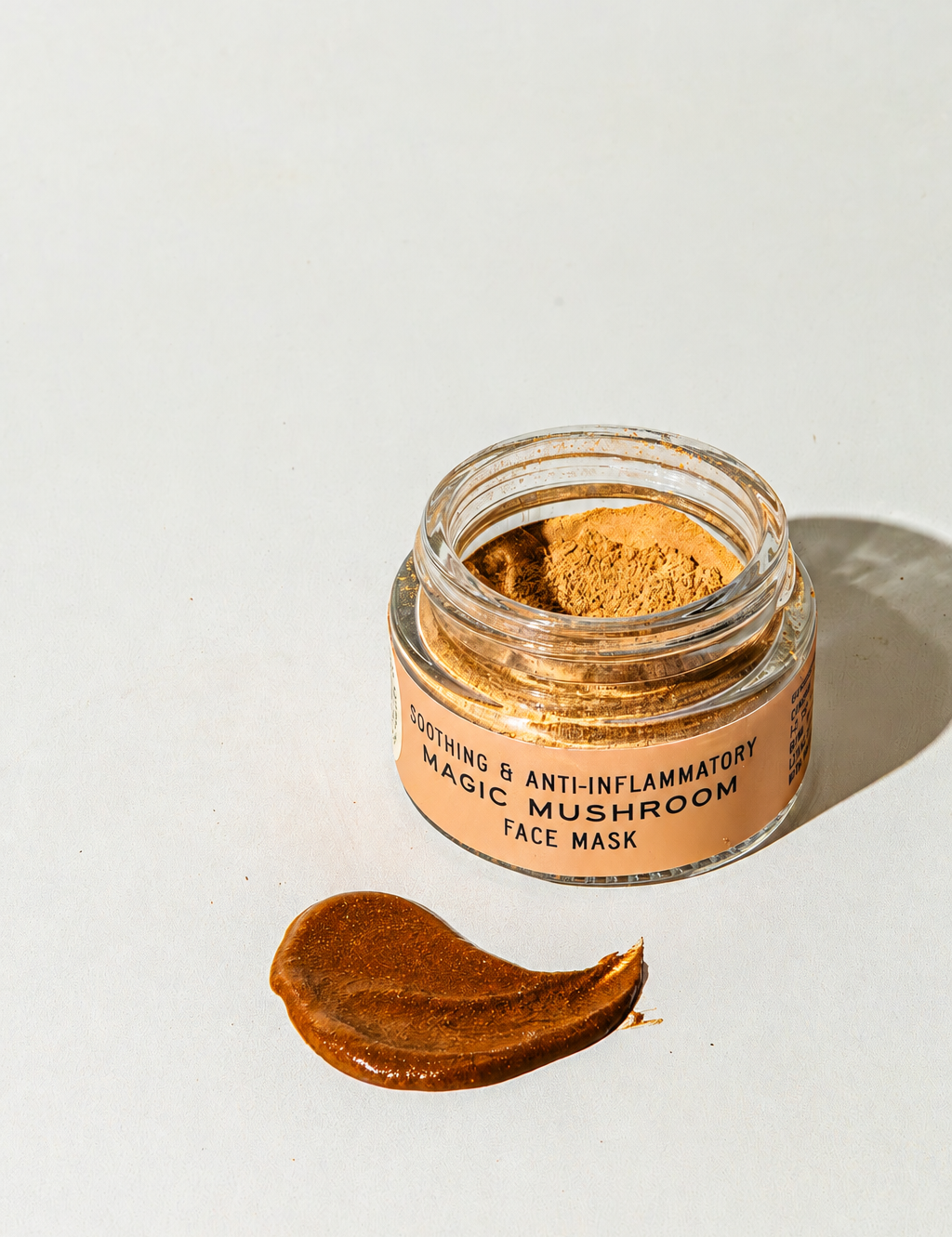 An open glass jar of Good Flower Farm Magic Mushroom Face Mask powder next to a smear of the product on a white surface, with a label reading Soothing & Anti-Inflammatory Magic Mushroom Face Mask.