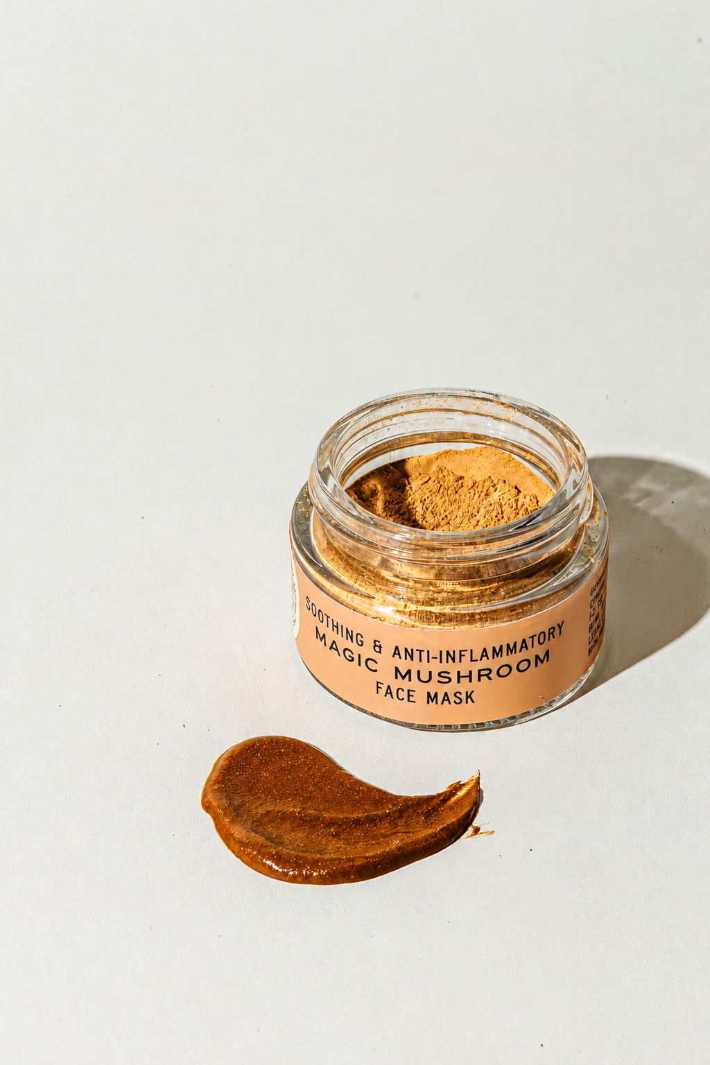 An open glass jar of Good Flower Farm Magic Mushroom Face Mask powder next to a smear of the product on a white surface, with a label reading Soothing &amp; Anti-Inflammatory Magic Mushroom Face Mask.