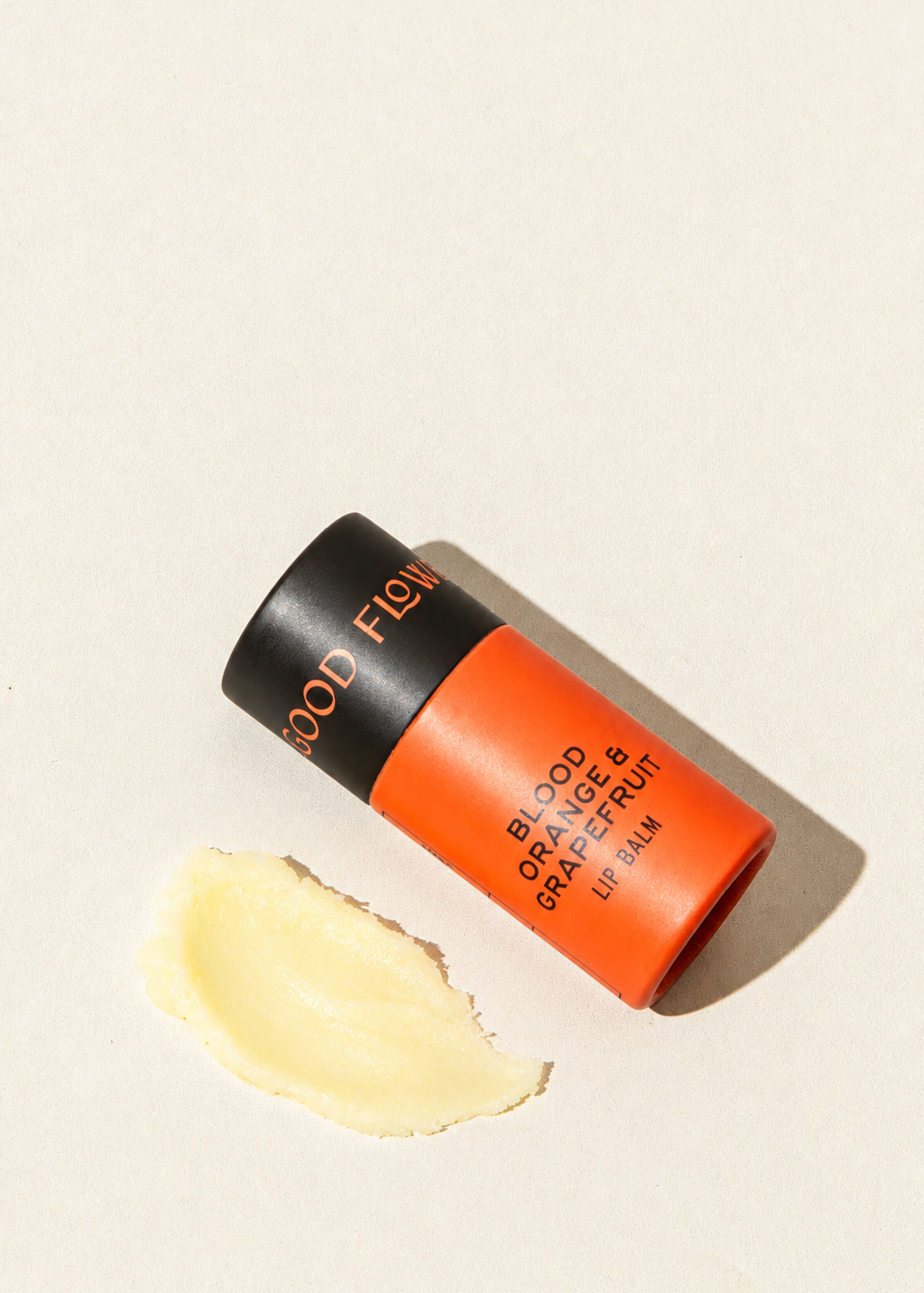 A tube of Good Flower Farm Blood Orange & Grapefruit Organic Lip Balm in eco-friendly packaging lies next to a small smear of the product.