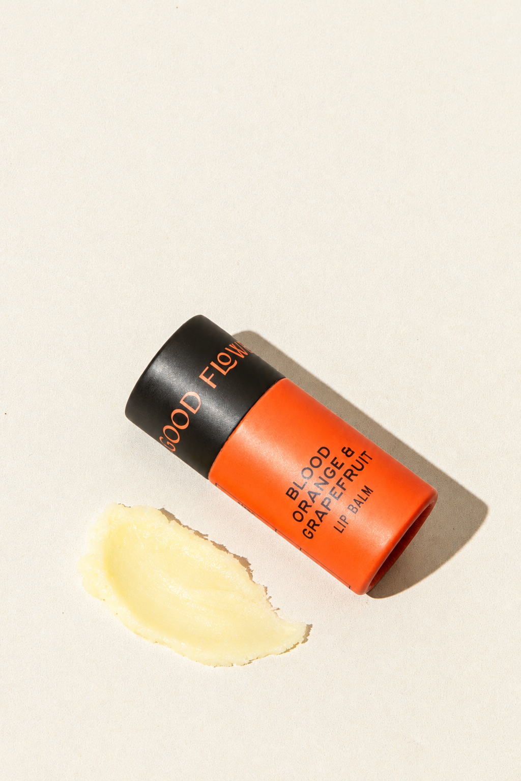 A tube of Good Flower Farm Blood Orange & Grapefruit Organic Lip Balm in eco-friendly packaging lies next to a small smear of the product.