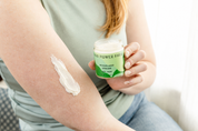 A person holds a glass jar of Good Flower Farm Woodland Dream Organic Body Cream while a dollop of the product is swiped onto their upper arm.