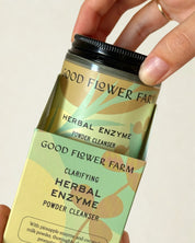 A hand takes a jar of Good Flower Farm Herbal Enzyme Powder Facial Cleanser from its green and yellow box, which showcases leafy designs and details about papaya enzymes on the vibrant packaging.
