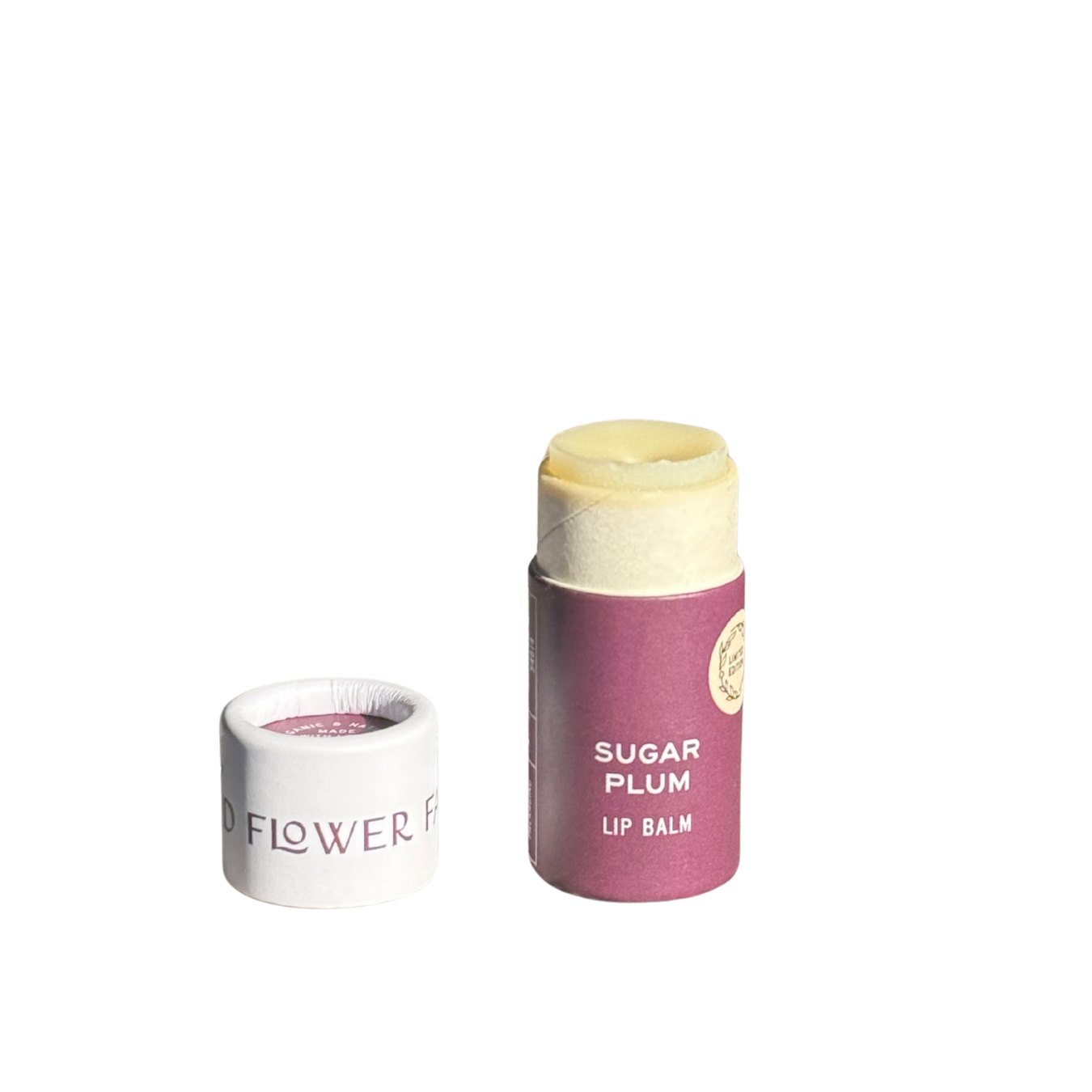 A SEASONAL LIMITED Holiday Lip Gift Set - NEW lip balm by Good Flower Farm with a purple label and biodegradable tube stands next to its white cap on a white background. The coconut oil balm is partially exposed above the tube.