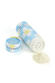 A cylindrical blue container from Good Flower Farm’s Good Baby Gift Set, filled with herbal baby powder, lies open as light-colored powder spills onto a white surface. The cap sits beside it and white and yellow flowers decorate the container.
